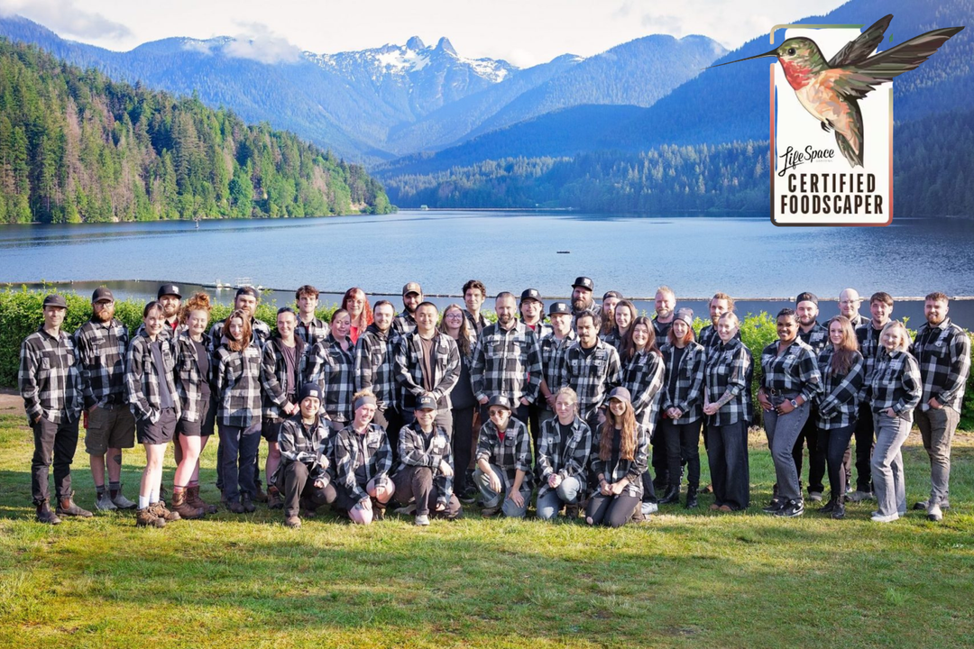 A large group of people posing outdoors in front of a mountain lake landscape, wearing matching black and white plaid flannel shirts. A badge in the top right reads 'The Space Certified Foodscaper' with a hummingbird graphic.