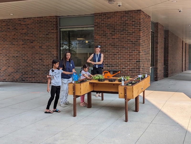 A family of four stands beside a wooden raised garden bed filled with plants and gardening tools, set outdoors against a brick building facade.