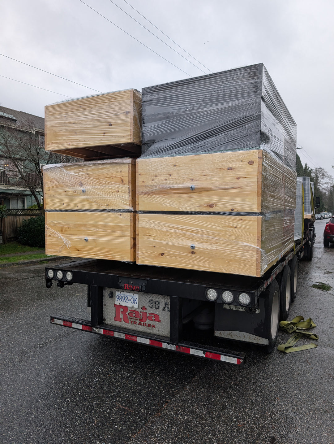 A flatbed truck loaded with large wooden crates and boxes wrapped in clear plastic, photographed outdoors on a wet street under an overcast sky. The truck bed has a visible 'Raja' license plate.
