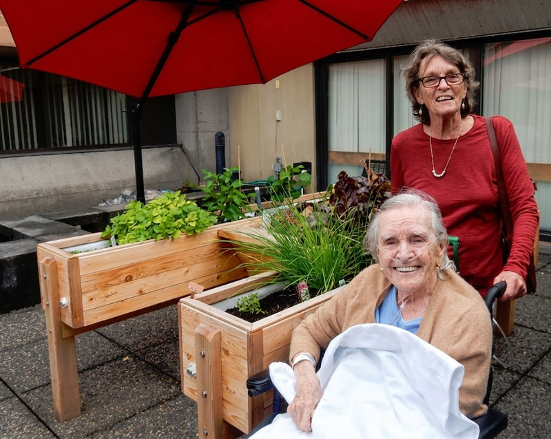 An elderly woman in a wheelchair wearing a tan cardigan and blue shirt smiles while holding a white blanket, with another woman in a red top standing behind her under a red patio umbrella beside wooden raised garden beds.